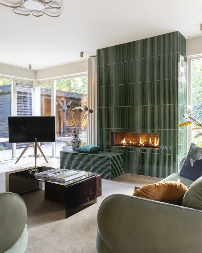Woonkamer met grote groen betegelde open haard, groene bank en bruin glazen salontafel.