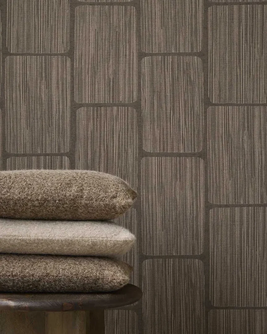 estapelde kussens in aardetinten op een houten kruk voor een wand met grafisch patroon.