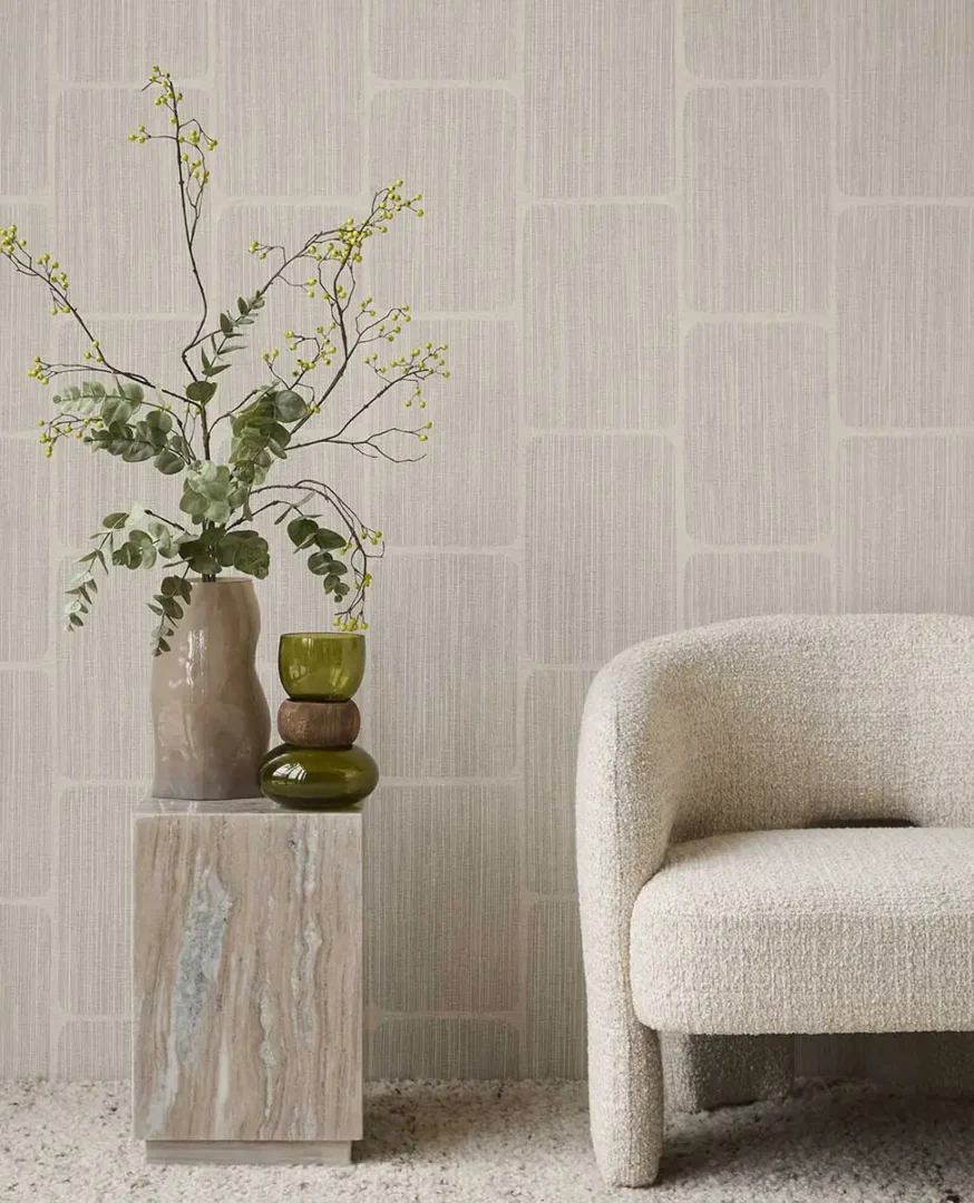 Stenen bijzettafel met keramische vaas en glazen decoratie naast een zachte fauteuil voor een wand met subtiel patroon.