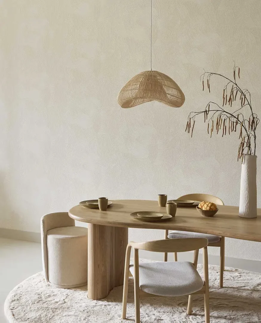 Ovale houten eettafel met keramisch servies en een witte vaas met takken onder een gevlochten hanglamp in een rustige, moderne eetruimte.