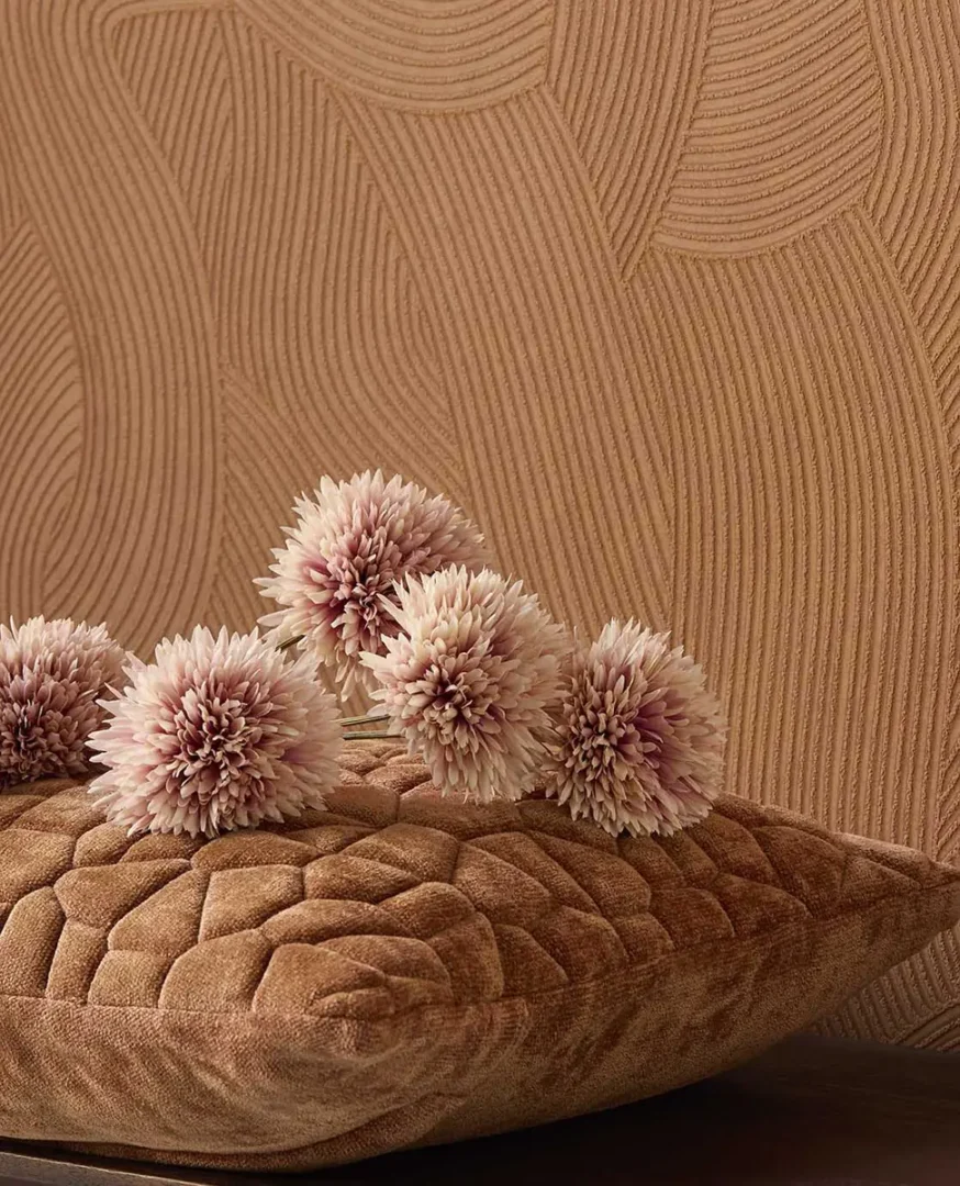 Bruin kussen met zachte kunstbloemen in crème‑ en rozetinten voor een wand met subtiel golvend reliëf.