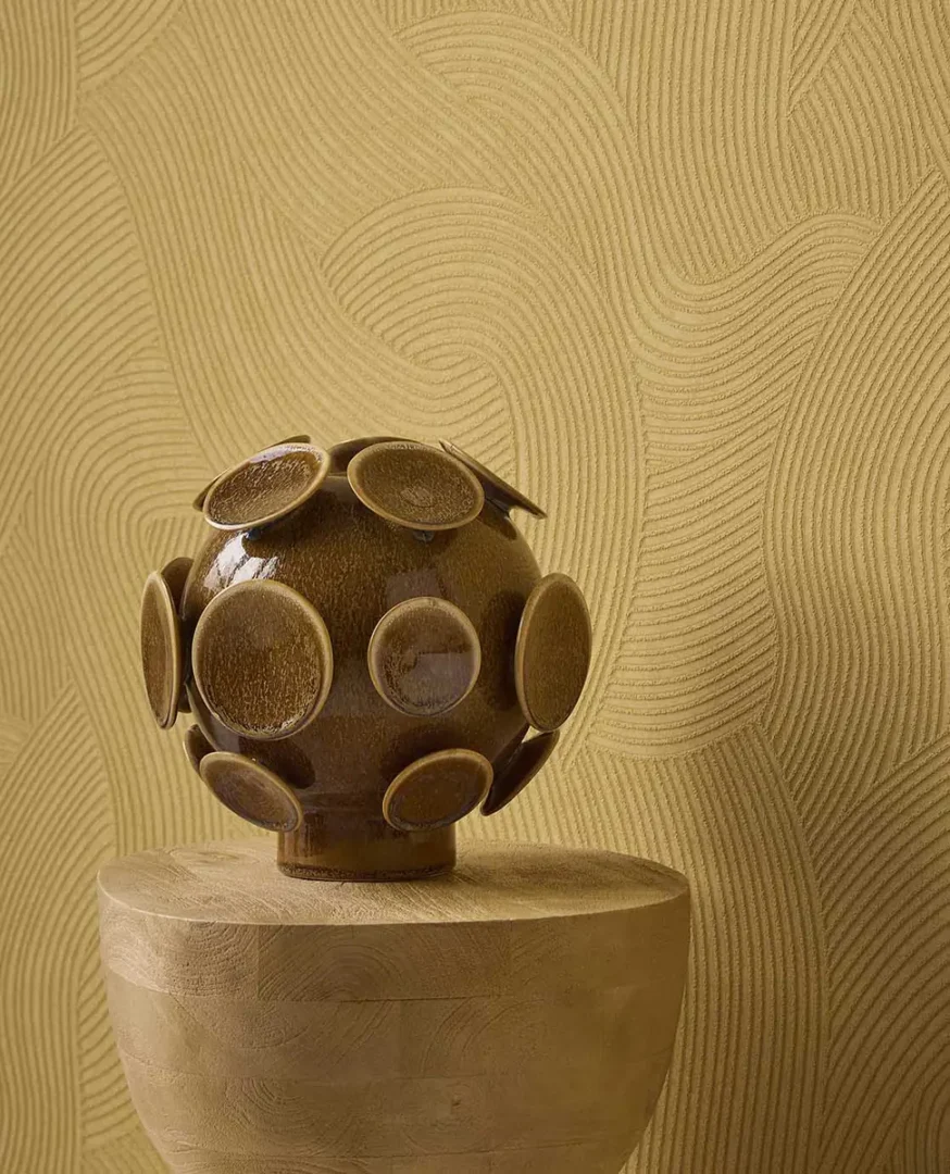 Glanzend bruine sculpturale bol op een houten sokkel voor een wand met subtiel golvend reliëf.