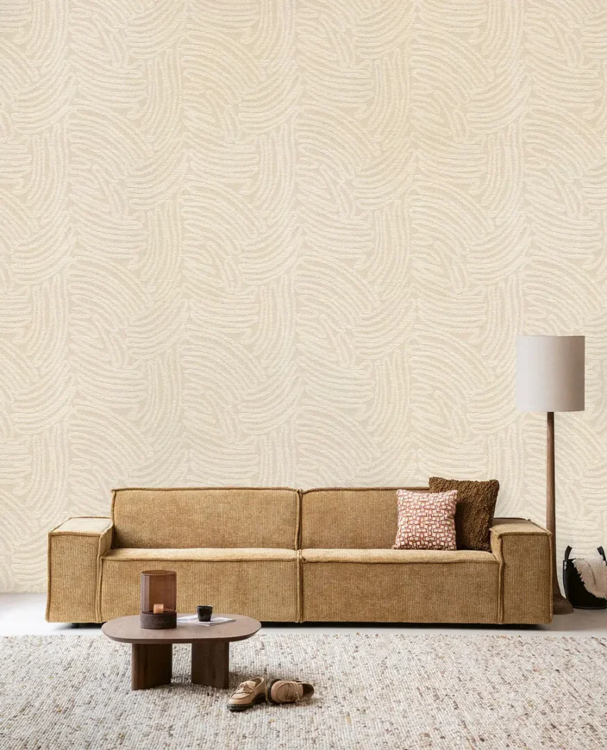 Tan corduroy bank met kussens, ronde houten salontafel en slippers op een licht vloerkleed voor een wand met subtiel lijnenspel.