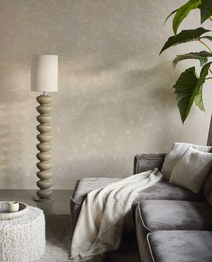 Grijze hoekbank met zachte plaid en sculpturale vloerlamp naast een ronde poef in een rustige, moderne woonkamer.