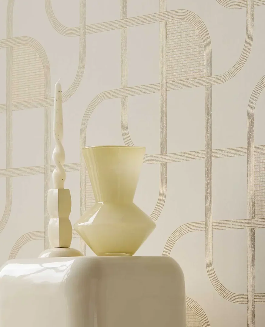 Gedraaide crème kaars en lichtgele geometrische vaas op een keramische sokkel voor een wand met subtiel lijnpatroon.”