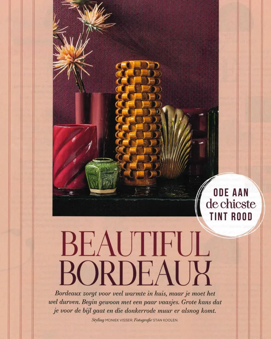 Decoratieve sculpturale bol en vazen in warme rood‑ en goudtinten tegen een rijke bordeaux achtergrond.