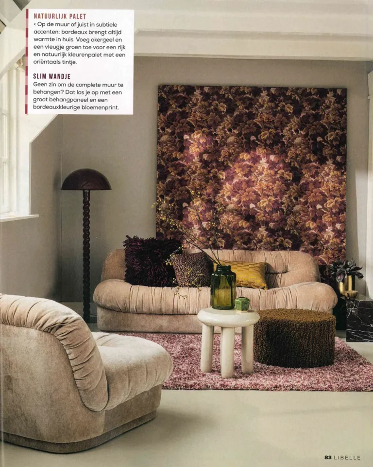 Beige bank en fauteuil met ronde vormen voor een bordeaux behangpaneel, met groene vazen op een witte salontafel en een mauve vloerkleed.