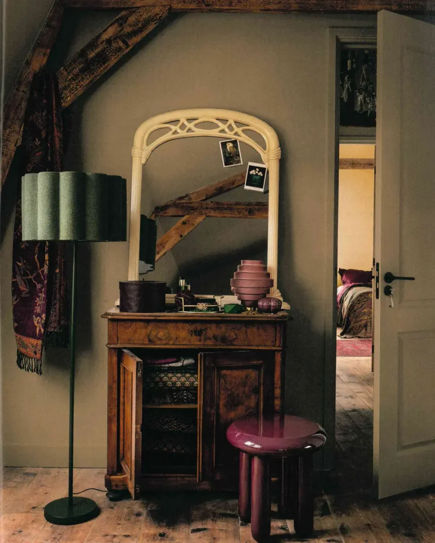 Houten kaptafel met spiegel, maroon kruk en groene vloerlamp in een warme, rustieke ruimte met zicht op een kleurrijke slaapkamer.