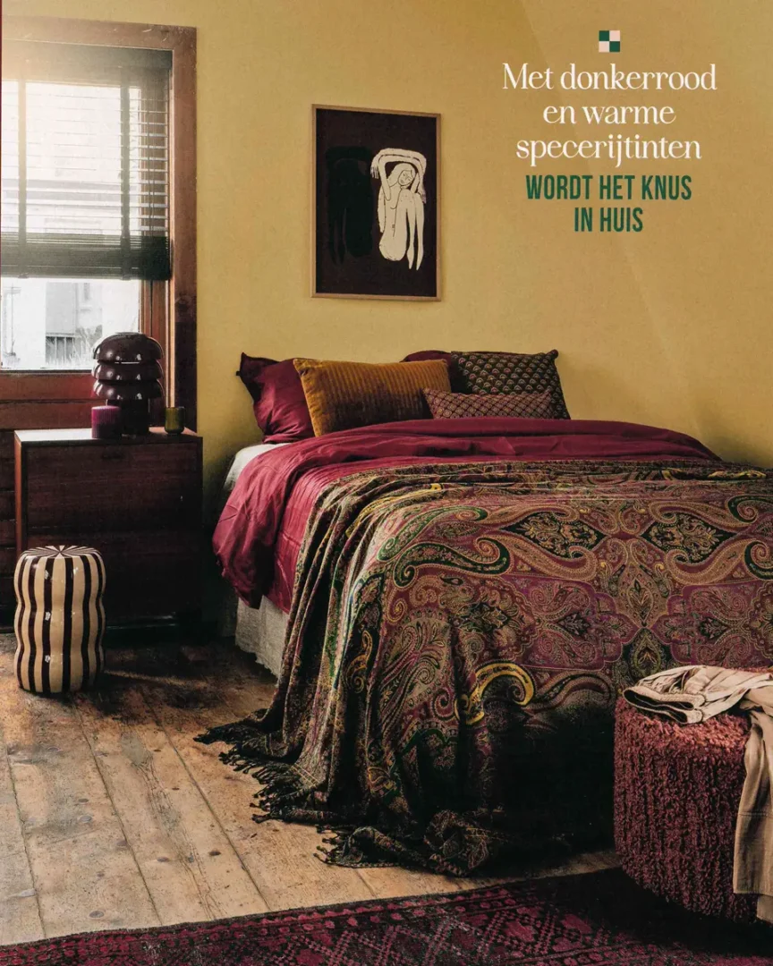 Slaapkamer met paisley sprei in diepe rood‑ en groentinten, bordeaux kussens en houten accenten in warm licht.