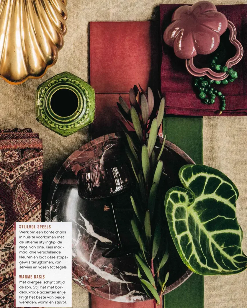 Flatlay met gouden schelp, groene vaas, roze schaaltje, bordeaux marmer en een groot groen blad op warme texturen.