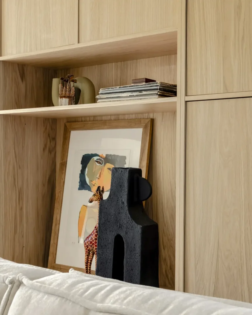 Detail van een lichte houten kast met open planken, waarop een kunstzinnige compositie te zien is. Een ingelijste illustratie van een vrouw met giraffe‑motief leunt tegen de achterwand, naast een zwarte sculptuur met architecturale vormen. Op de bovenste plank liggen tijdschriften en keramiek, wat een rustige, natuurlijke sfeer creëert in het interieur.