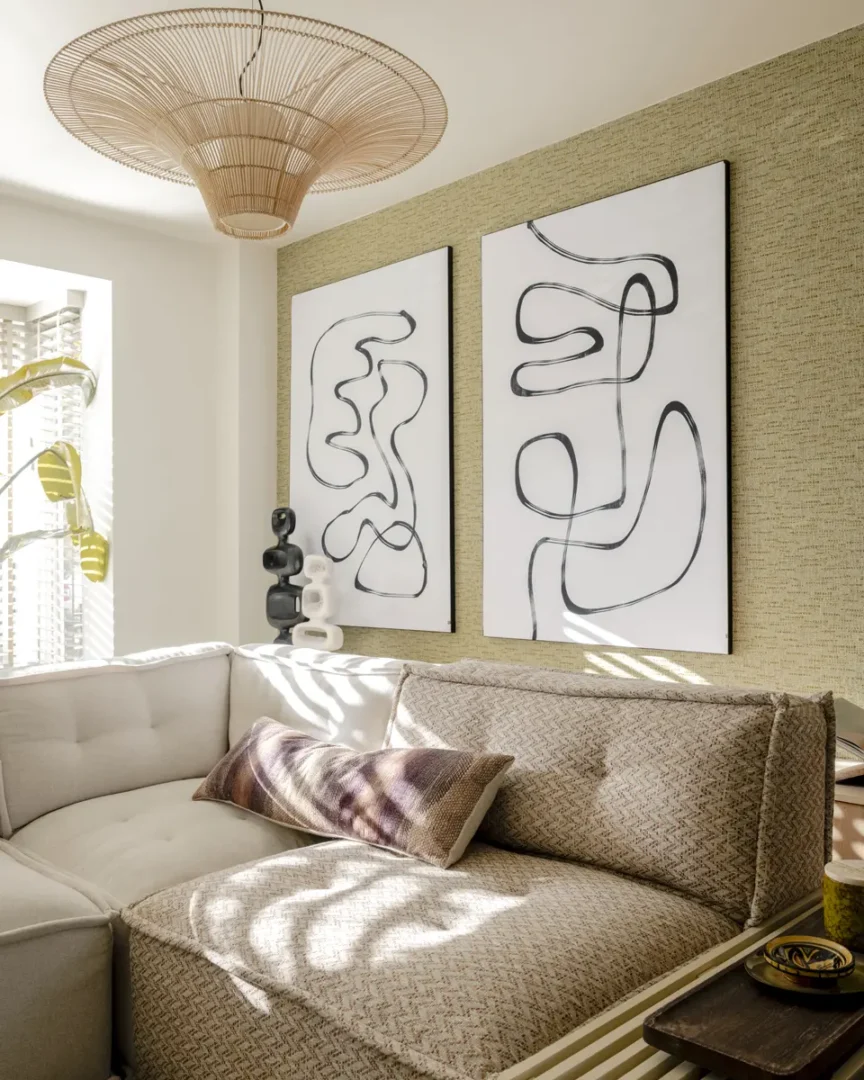 Zonnige woonkamer met een licht hoekbank en twee abstracte zwart‑witte kunstwerken aan de muur. De ruimte ademt rust en textuur, met een rotan plafondlamp, een sculpturaal zwart beeld en natuurlijke accenten in beige en groen. Zachte schaduwen vallen over de bank en kussens, wat een serene, uitnodigende sfeer creëert.