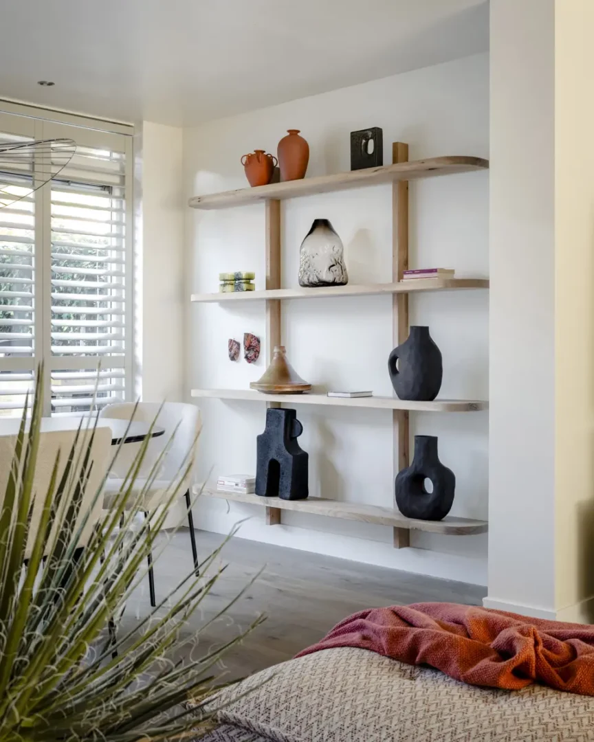 Een lichte woonkamer met een open houten wandrek vol keramiek en sculpturale objecten in aardetinten. Op de planken staan terracotta vazen, kunstzinnige vormen en glaswerk, gecombineerd met boeken en kleine kunstwerken. Voor het rek staat een witte stoel bij het raam met shutters, terwijl een plaid en kussen in zachte tinten de ruimte warmte geven.