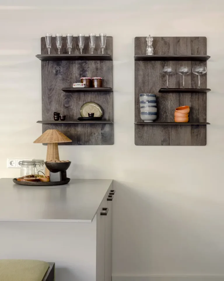 Twee wandplanken van donker hout met glazen, keramiek en vintage servies in een lichte keukenhoek. Op het grijze werkblad staat een rotan tafellamp en een schaal met accessoires. De compositie straalt rust, textuur en natuurlijke harmonie uit.