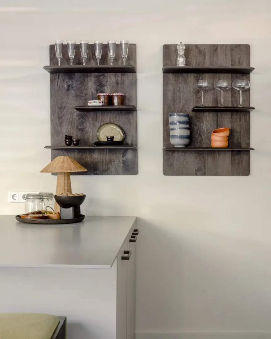 Twee wandplanken van donker hout met glazen, keramiek en vintage servies in een lichte keukenhoek. Op het grijze werkblad staat een rotan tafellamp en een schaal met accessoires. De compositie straalt rust, textuur en natuurlijke harmonie uit.