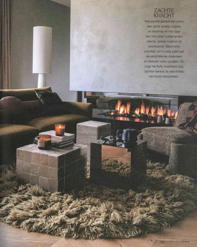 Woonkamer met brandende haard, fluffy vloerkleed, natuurstenen salontafels en een olijfgroene bank in zachte aardetinten.