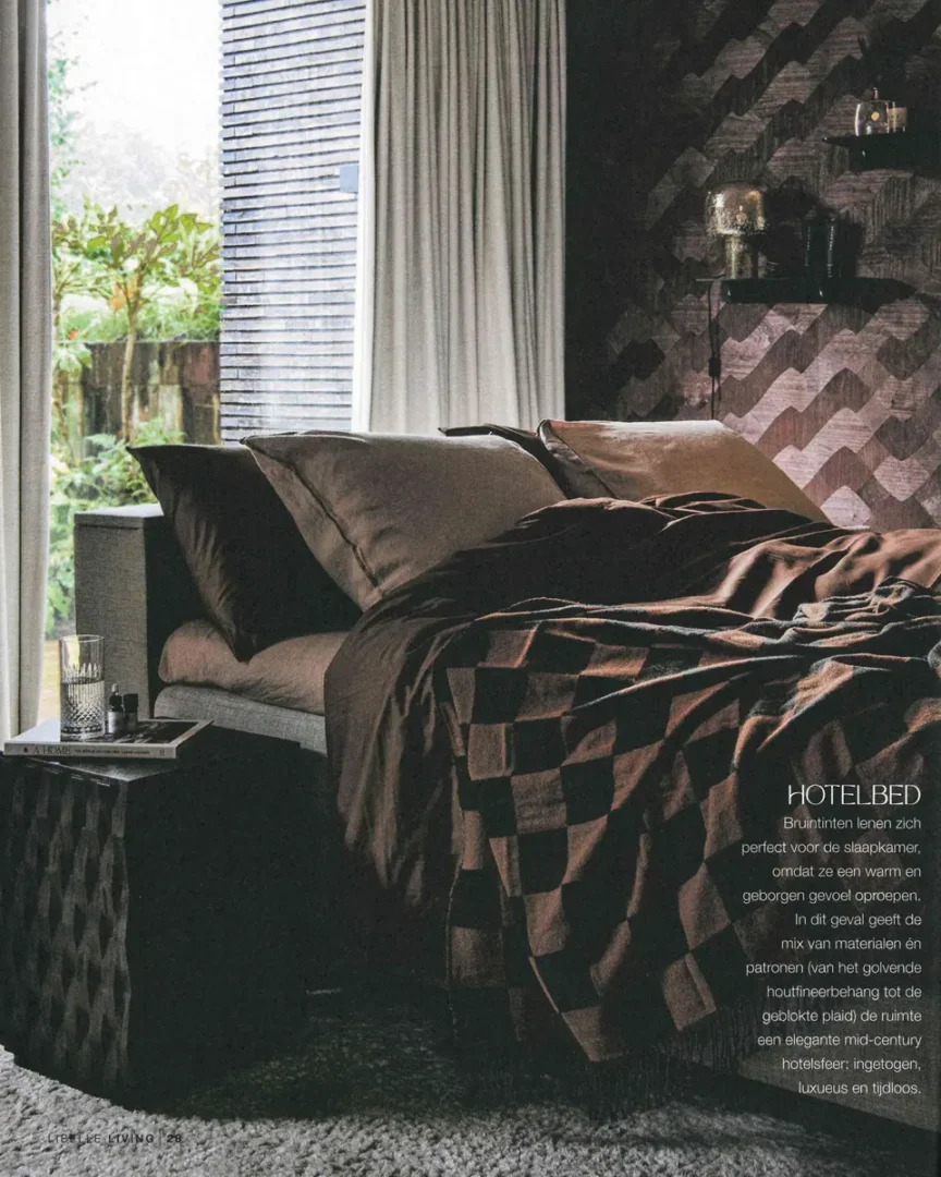 Slaapkamer met bruintinten, een bed met geblokte plaid, donker nachtkastje en een geometrische houtwand met decoratieve items.