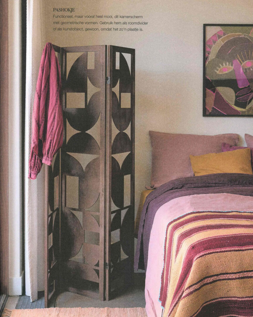 Een moderne woonkamer met een open vakkenkast van licht hout, gevuld met keramiek en vazen in roze en terracotta tinten. De kast fungeert als roomdivider tussen eet‑ en zithoek. Op de achtergrond een beige bank met kussens en plaid in warme kleuren, een donker houten plafond en grote ramen die natuurlijk licht binnenlaten. De sfeer is zacht, eigentijds en harmonieus.