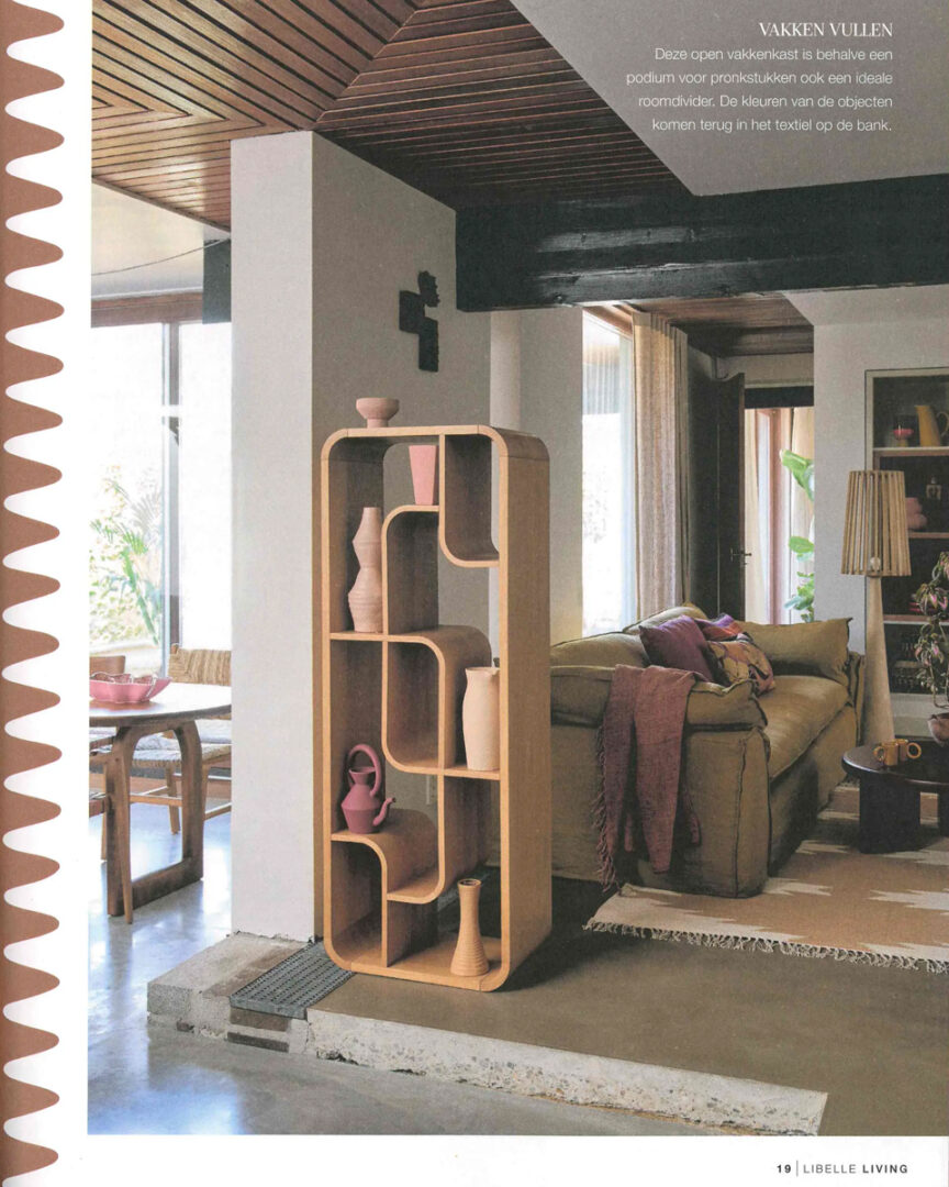 Een moderne woonkamer met een open vakkenkast van licht hout, gevuld met keramiek en vazen in roze en terracotta tinten. De kast fungeert als roomdivider tussen eet‑ en zithoek. Op de achtergrond een beige bank met kussens en plaid in warme kleuren, een donker houten plafond en grote ramen die natuurlijk licht binnenlaten. De sfeer is zacht, eigentijds en harmonieus.