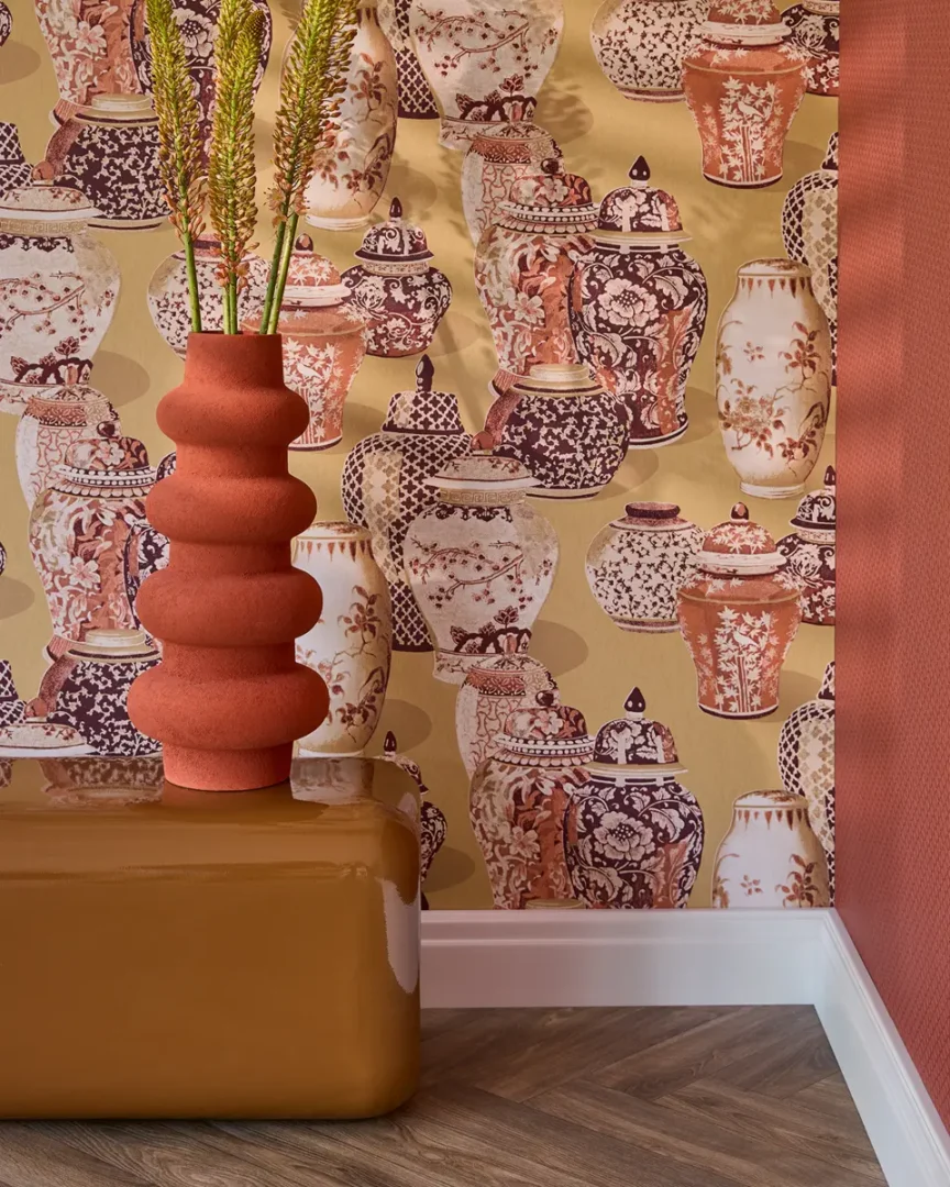 Interieurhoek met behang vol klassieke vazen in warme tinten, een terracotta sculptuurvaas en een glanzend okerkleurig meubel.