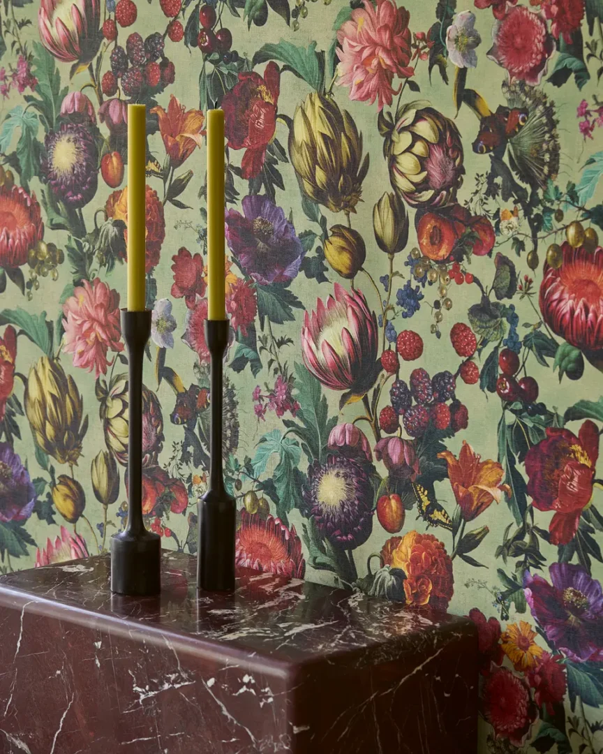 Interieurhoek met marmeren console, twee zwarte kandelaars met gele kaarsen en een wand vol kleurrijke bloemenprint.”