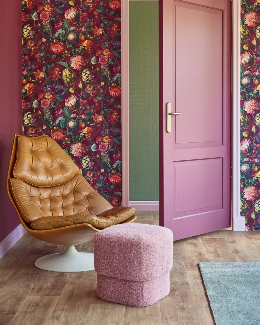 Interieurhoek met cognackleurige fauteuil, roze poef en een wand met rijk bloemenbehang in warme tinten.