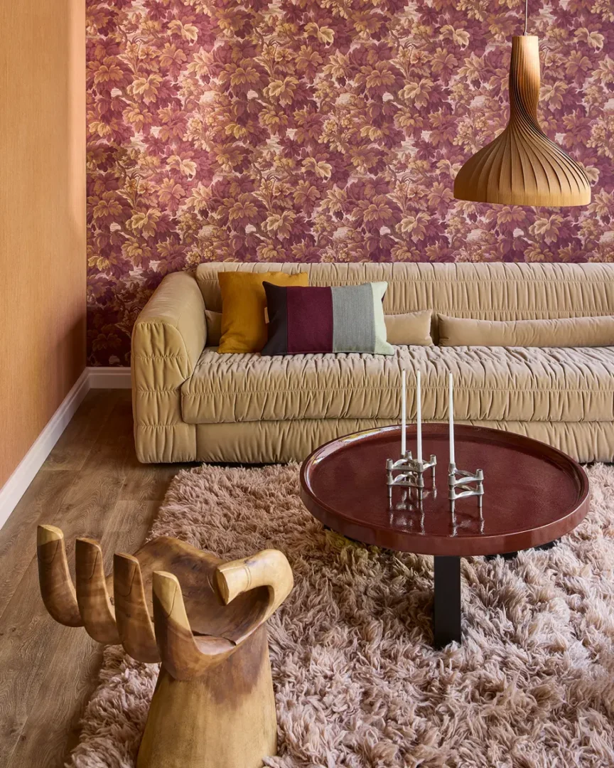 Woonkamer met beige ruchebank, ronde tafel, houten handstoel en bloemrijk behang in roze‑gouden tinten.