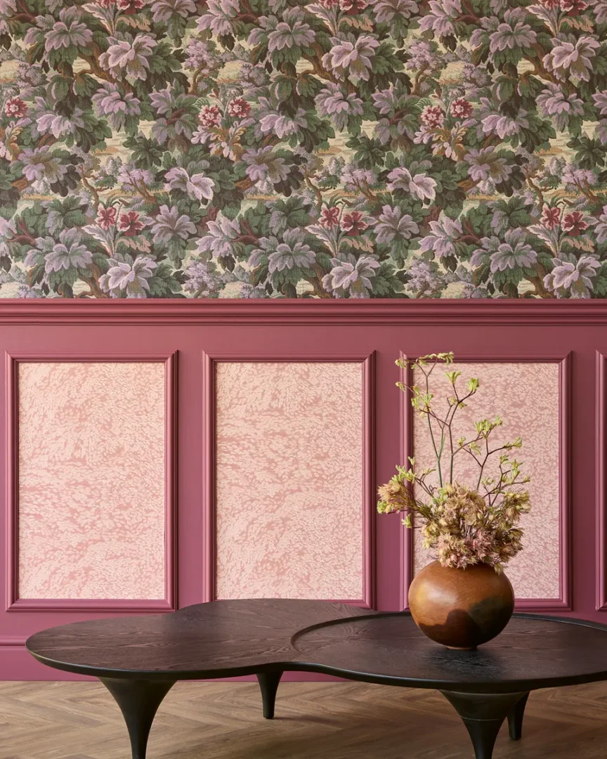 Interieurhoek met bloemrijk behang, roze lambrisering, donkere organische tafels en een aardse vaas met gedroogde bloemen.