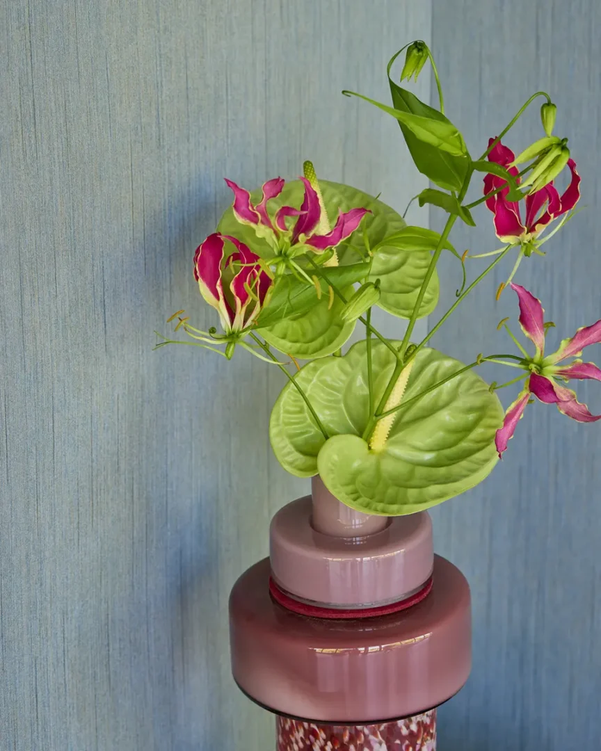 Keramische vaas in roze en roodtinten met groene bladeren en roze bloemen tegen een blauwgrijze wand.
