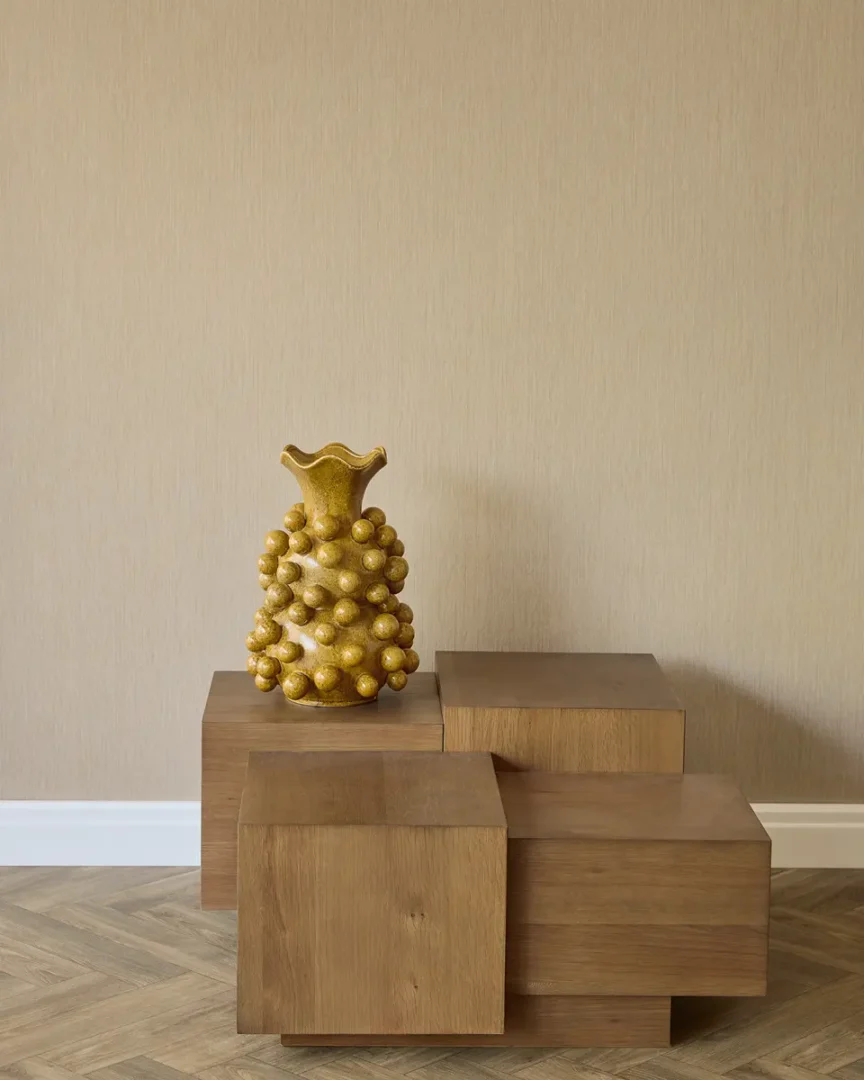 Minimalistische interieurhoek met gouden sculptuurvaas op drie houten blokken tegen een beige wand en visgraatvloer.