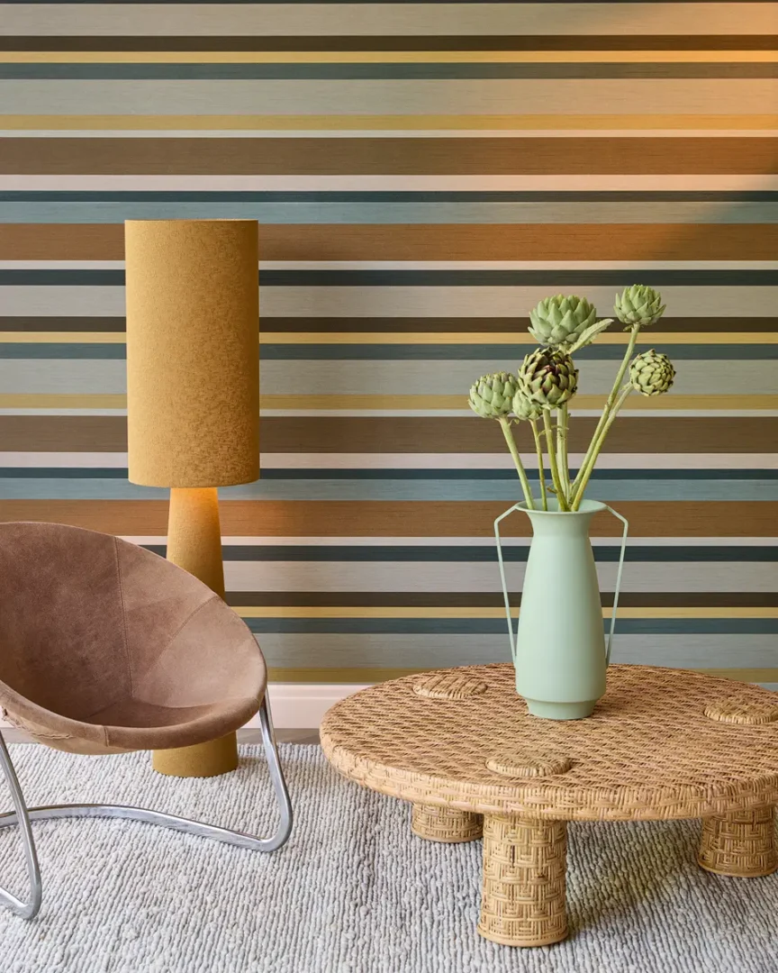 Interieurhoek met gestreept behang in aardse tinten, rotan tafel, mintgroene vaas met artisjokken, leren stoel en okergele lamp.