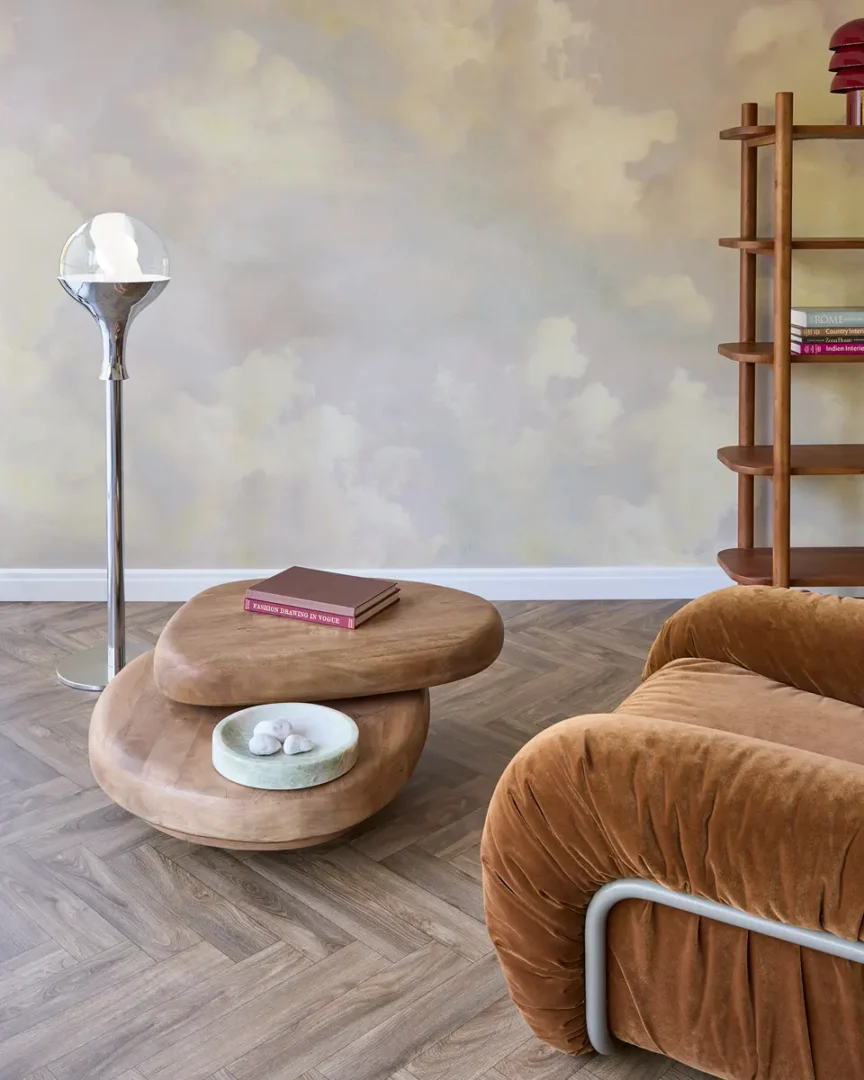 Woonkamer met fluwelen fauteuil, sculpturale houten salontafel, chromen vloerlamp en wolkenachtig behang in zachte tinten.