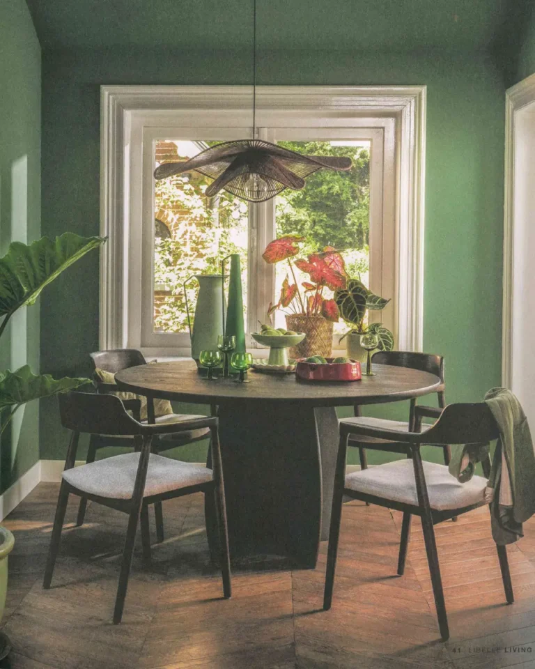 Eetkamer met groene muren, ronde houten tafel, vier stoelen en planten bij een raam dat uitkijkt op een groene tuin.