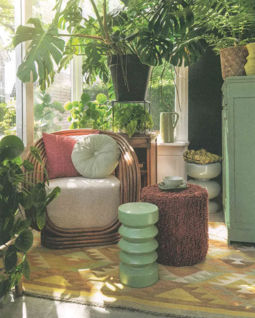Zonnige hoek met rotan fauteuil, roze en groene kussens, veel kamerplanten en een zacht geometrisch vloerkleed.