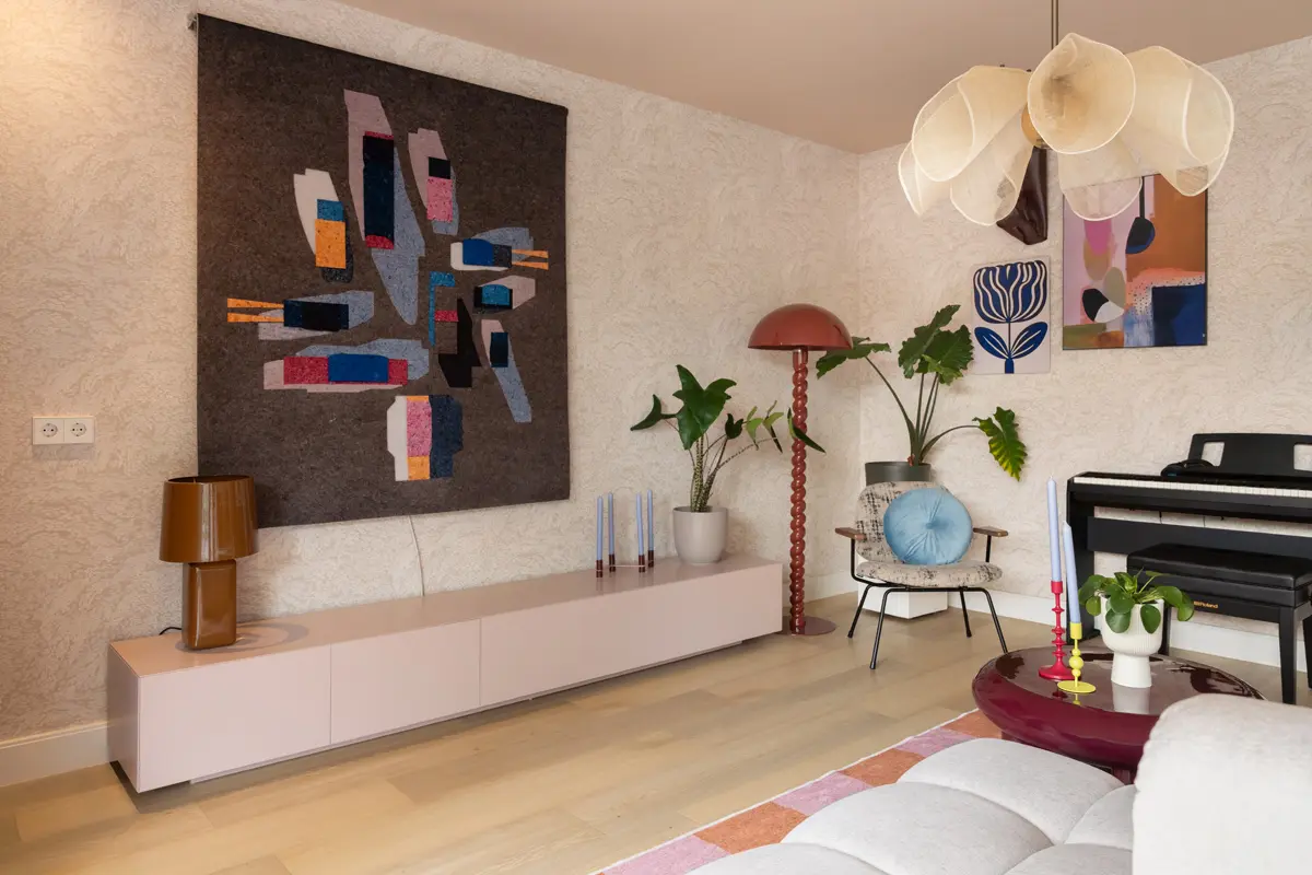 Moderne woonkamer met abstract wandkleed, rode sculpturale lamp, designstoel met blauw kussen, piano en zachte tinten in een lichte setting.