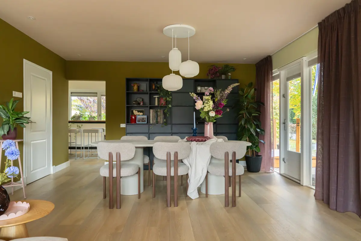 Eetkamer met olijfgroene wand, ronde tafel met zes beklede stoelen, zwarte kastwand met planten en boeken, en grote ramen met zicht op de tuin.