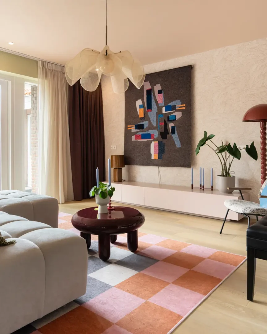 Moderne woonkamer met lichtgrijze bank, kleurrijk wandkleed, roze‑oranje vloerkleed en ronde bordeauxrode tafel met groene vaas.