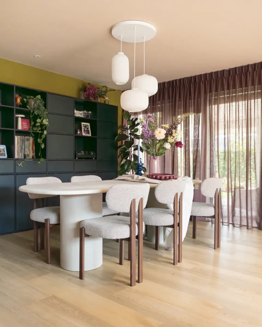 Eetkamer met ronde tafel en zes beklede stoelen, zwarte kastwand met planten en boeken, en grote ramen met mauve gordijnen die zacht licht binnenlaten.