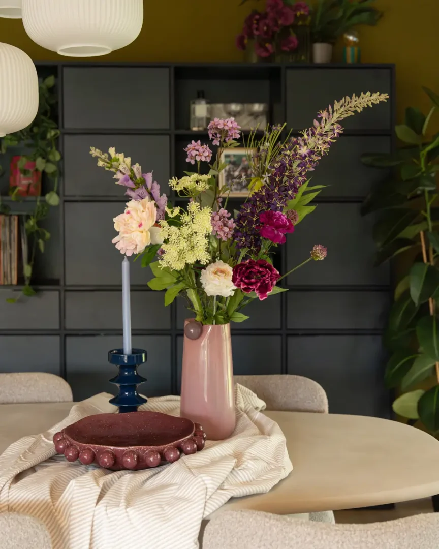 Close‑up van een eettafel met roze keramische vaas vol kleurrijke bloemen, blauwe kandelaar, ronde schaal met kralenrand en zachte stoffen ondergrond.