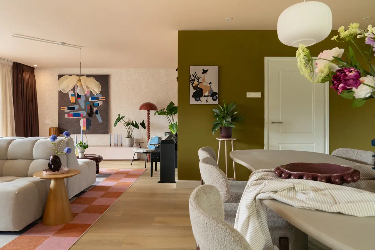 Open woon‑ en eetruimte met olijfgroene wand, lichtgrijze bank, kleurrijk wandkleed, ronde tafel met bloemen en zachte tinten in een moderne setting.