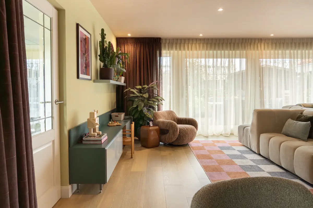 Lichte woonkamer met grote ramen en transparante gordijnen, beige bank, ronde bruine fauteuil, groene wand met kast en planten, en roze‑oranje vloerkleed.