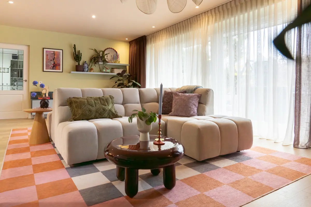 Lichte woonkamer met grote ramen en transparante gordijnen, beige modulaire bank, ronde bruine tafel met witte vaas, en roze‑oranje vloerkleed op houten vloer.