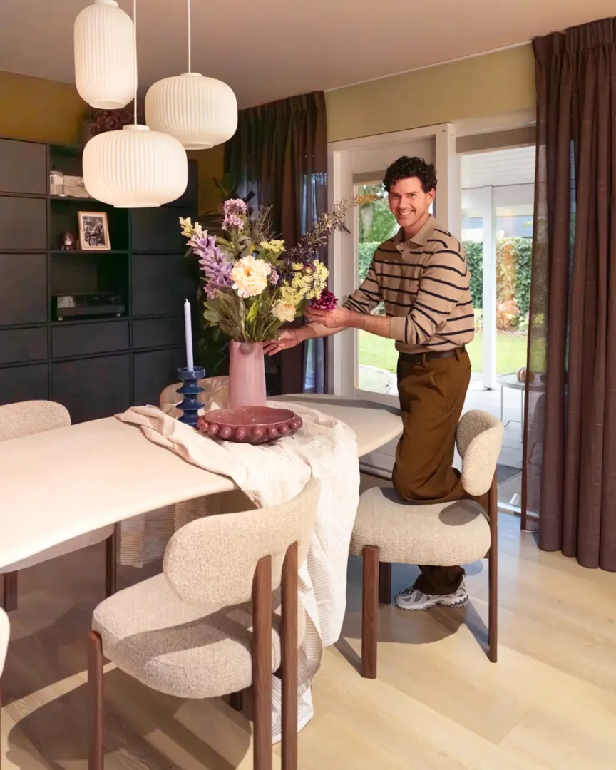 Eetkamer met ronde tafel, beige stoelen en roze vaas vol kleurrijke bloemen, terwijl een persoon met gestreepte trui de compositie verzorgt onder drie witte hanglampen.